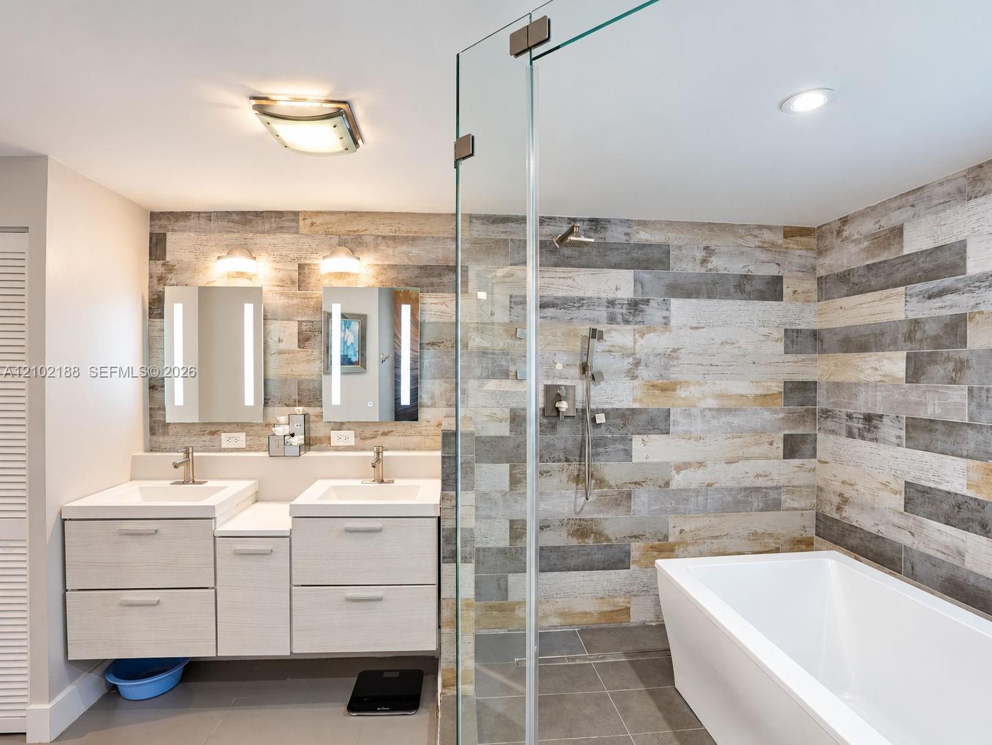 Dual-sink vanity with rectangular basins, integrated LED-lit mirrors, a glass-enclosed walk-in shower, freestanding soaking tub, and multi-toned wood-finish wall tile