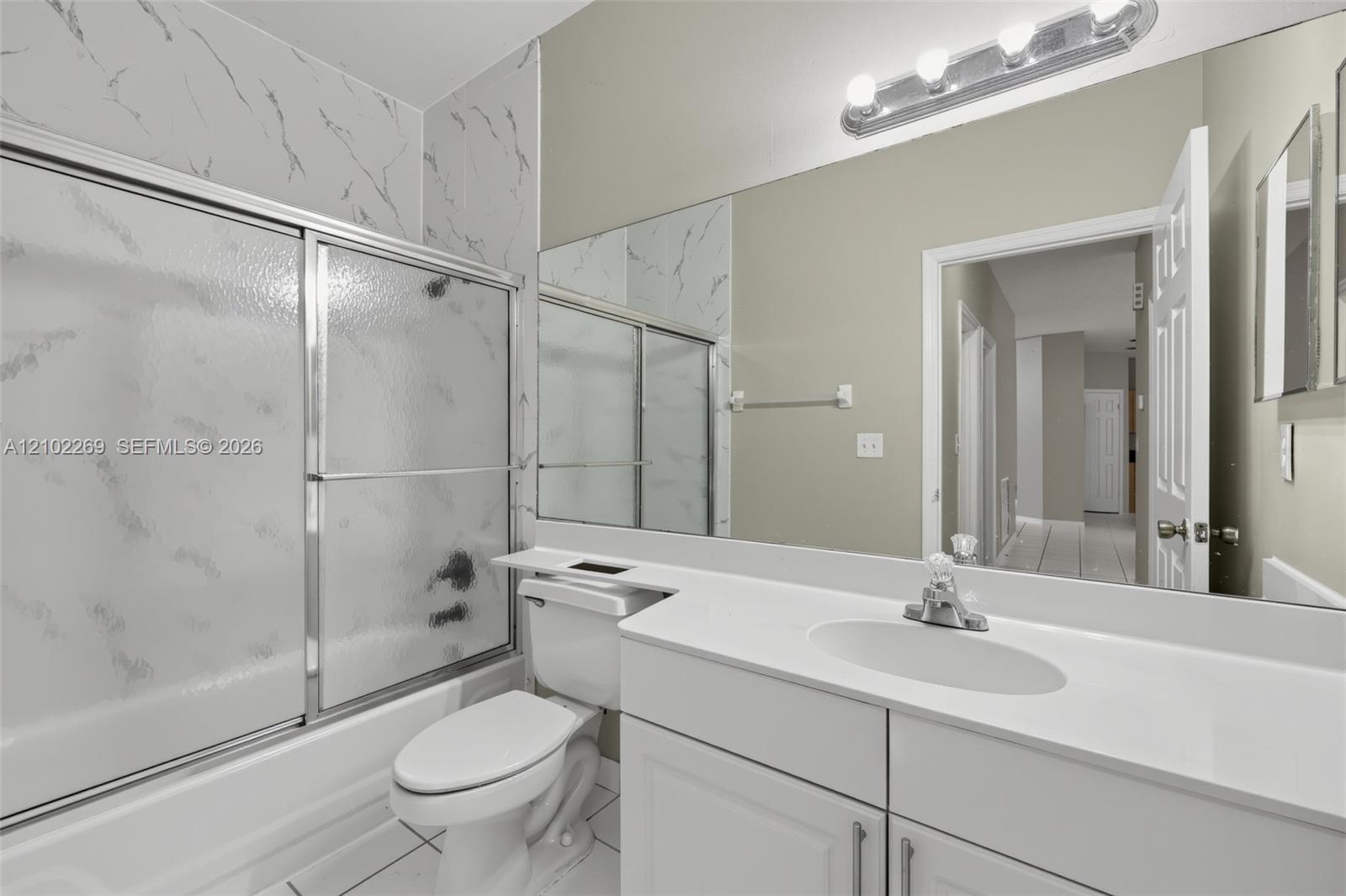 Bathroom featuring a white vanity with a solid surface countertop, an integrated oval sink, a full-width wall mirror, and a tub-shower combination with patterned wall surround and sliding glass doors Bathroom featuring a white vanity with a solid surface countertop, an integrated oval sink, a full-width wall mirror, and a tub-shower combination with patterned wall surround and sliding glass doors