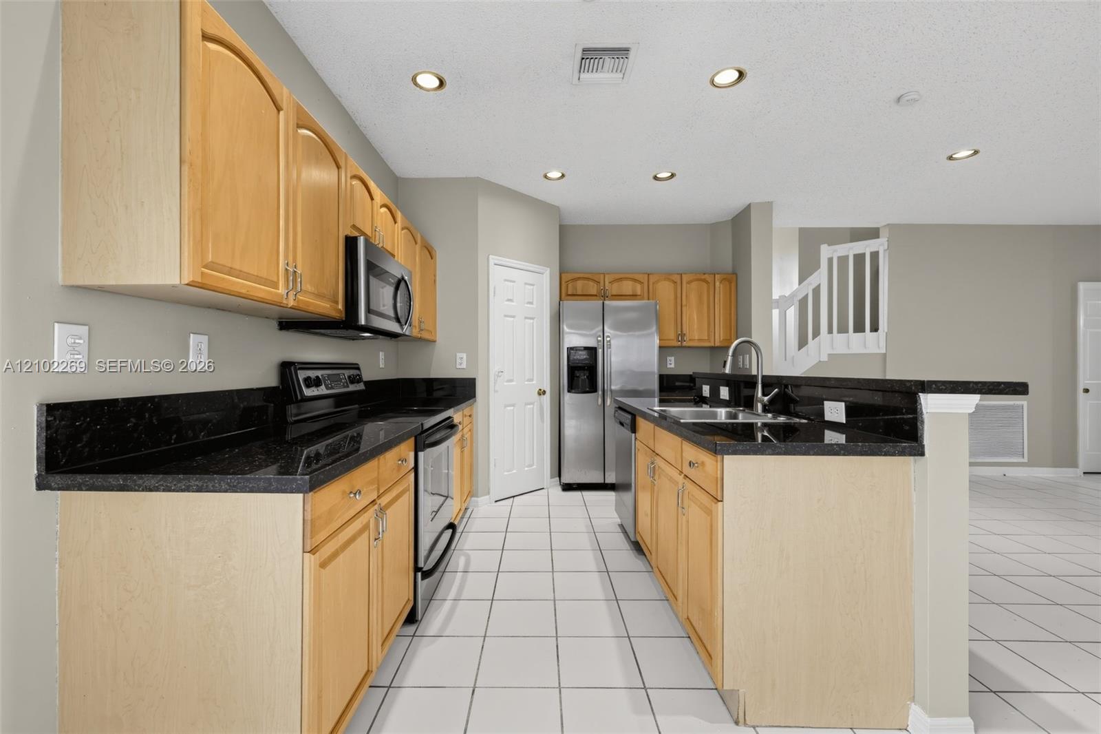 Kitchen featuring light wood cabinetry, black countertops, stainless steel appliances, recessed ceiling lighting, and ceramic tile flooring