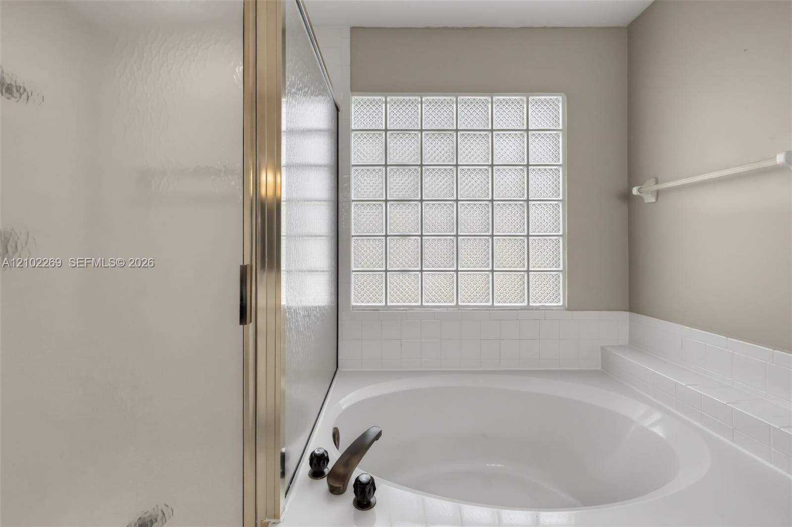 Oval soaking tub with a white tile surround, glass block window, and metal-finished hardware