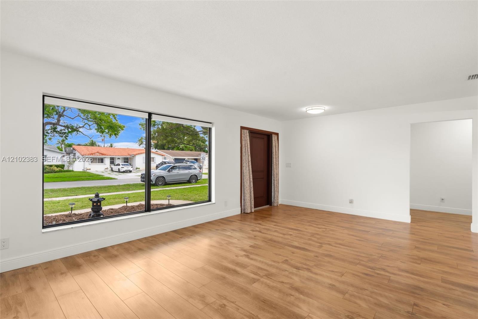 Interior space featuring wood-finish flooring, a large picture window, a solid-panel door, and a ceiling-mounted light fixture Interior space featuring wood-finish flooring, a large picture window, a solid-panel door, and a ceiling-mounted light fixture
