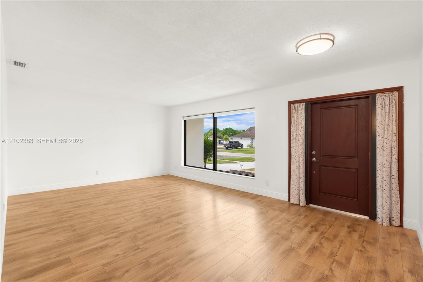 Interior space featuring wood-finish flooring, a large picture window, a solid wood-panel entry door, and a flush-mount ceiling light fixture Interior space featuring wood-finish flooring, a large picture window, a solid wood-panel entry door, and a flush-mount ceiling light fixture