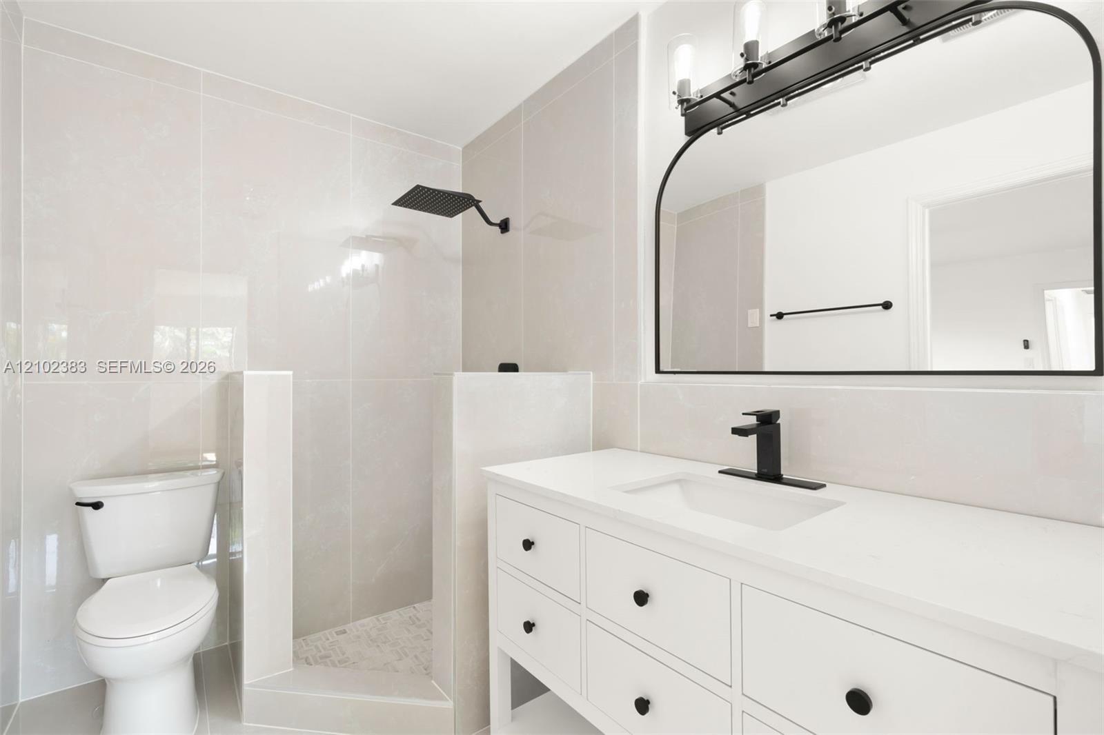 Primary bathroom featuring a white vanity with a quartz countertop, matte black faucet, wall-mounted rain shower head, arched vanity mirror, and light-toned wall tile