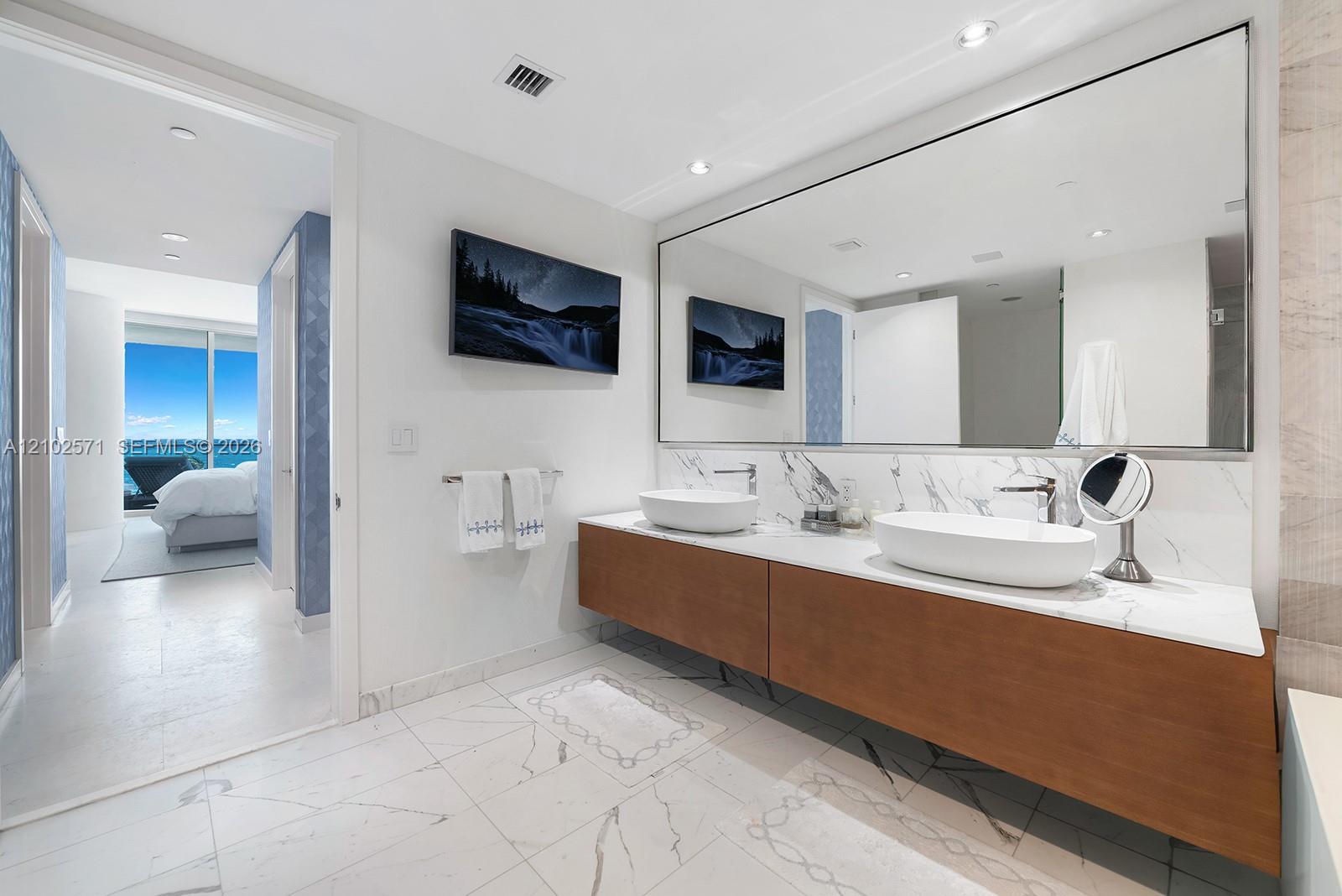 Floating wood-finish vanity with dual vessel sinks, stone-surface countertop, floor-to-ceiling mirror, and stone-finish tile flooring