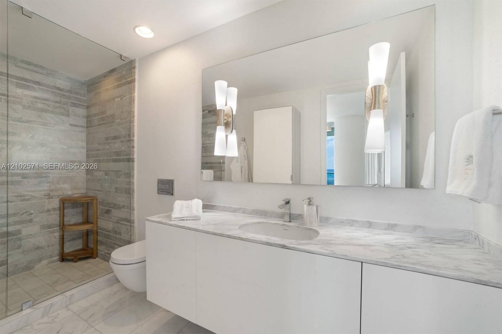 Floating vanity with integrated sink, stone-surface countertop, walk-in glass-enclosed shower featuring horizontal stone-tiled walls, wall-mounted toilet, and dual decorative light fixtures