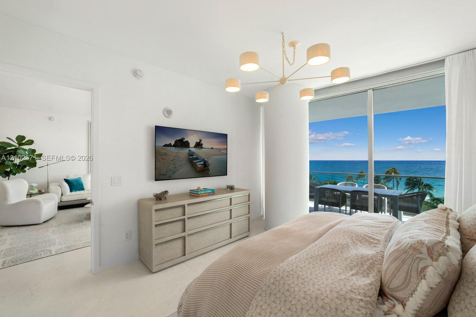 Full-height glass sliding doors, modern six-arm drum chandelier, and polished neutral-toned flooring with ocean backdrop and private balcony