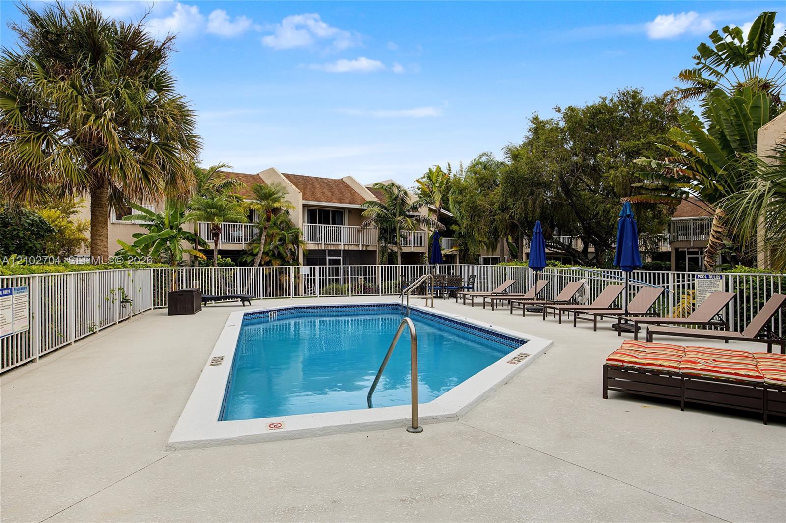 Community swimming pool featuring a concrete deck, white metal perimeter fencing, and mature palm tree landscaping