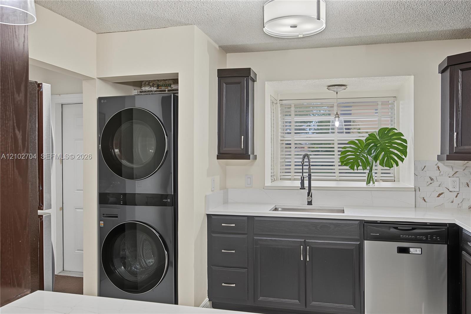 Dark-toned cabinetry, white solid-surface countertops, integrated stackable laundry unit, stainless steel dishwasher, and recessed window with blinds