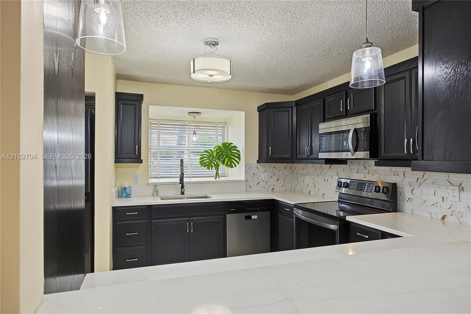 Kitchen featuring dark shaker cabinetry, light-toned stone-finish countertops, stainless steel appliances, patterned tile backsplash, and two glass pendant lights