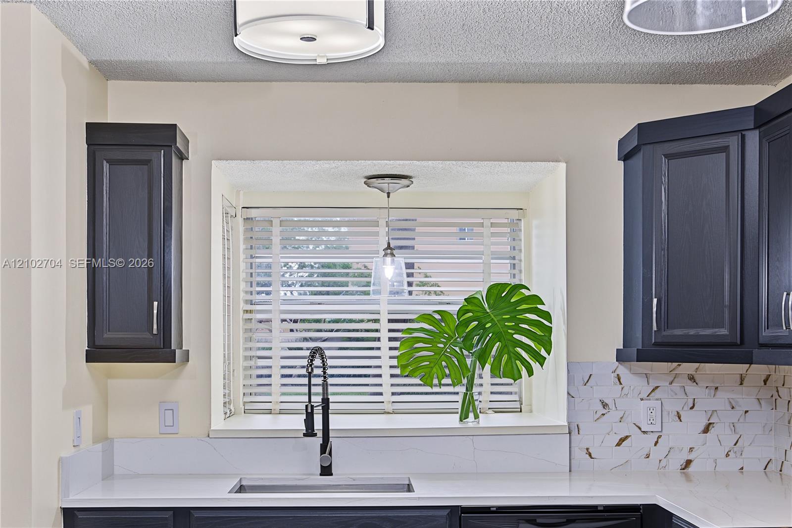 Kitchen featuring dark cabinetry, solid-surface countertops, decorative stone backsplash, undermount sink with a high-arch pull-down faucet, and a recessed window nook