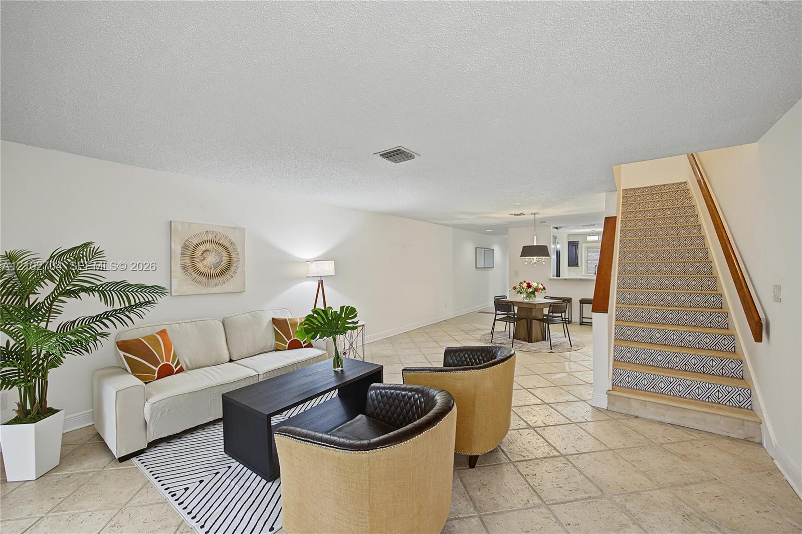 Open-concept interior featuring neutral tile flooring, a staircase with patterned riser detailing, a wooden handrail, and a pass-through breakfast bar