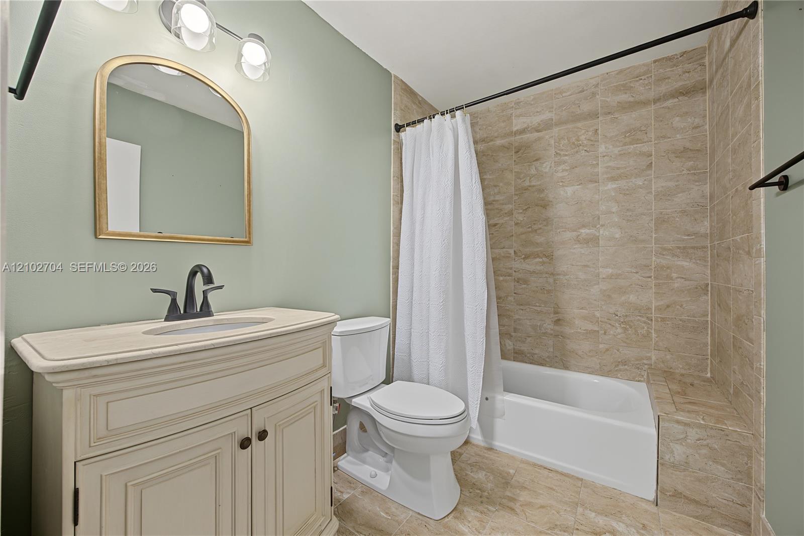 Bathroom featuring a vanity with a stone countertop and under-mount sink, matte black faucet hardware, arched wall mirror, light fixture with three glass shades, and a tile-surround shower and tub combination