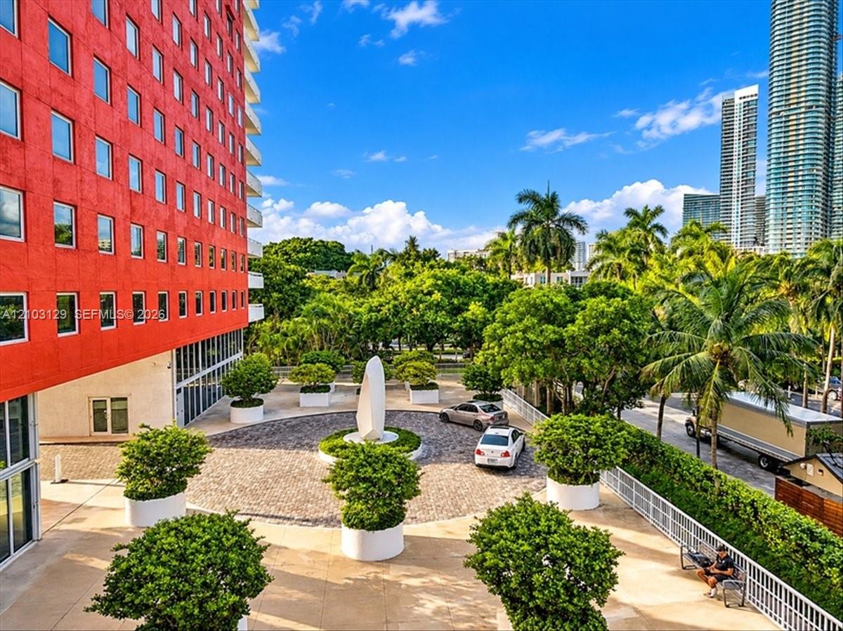 High-rise exterior featuring vibrant red concrete facade, circular cobblestone motor court, central sculptural focal point, and landscaped planters with integrated greenery