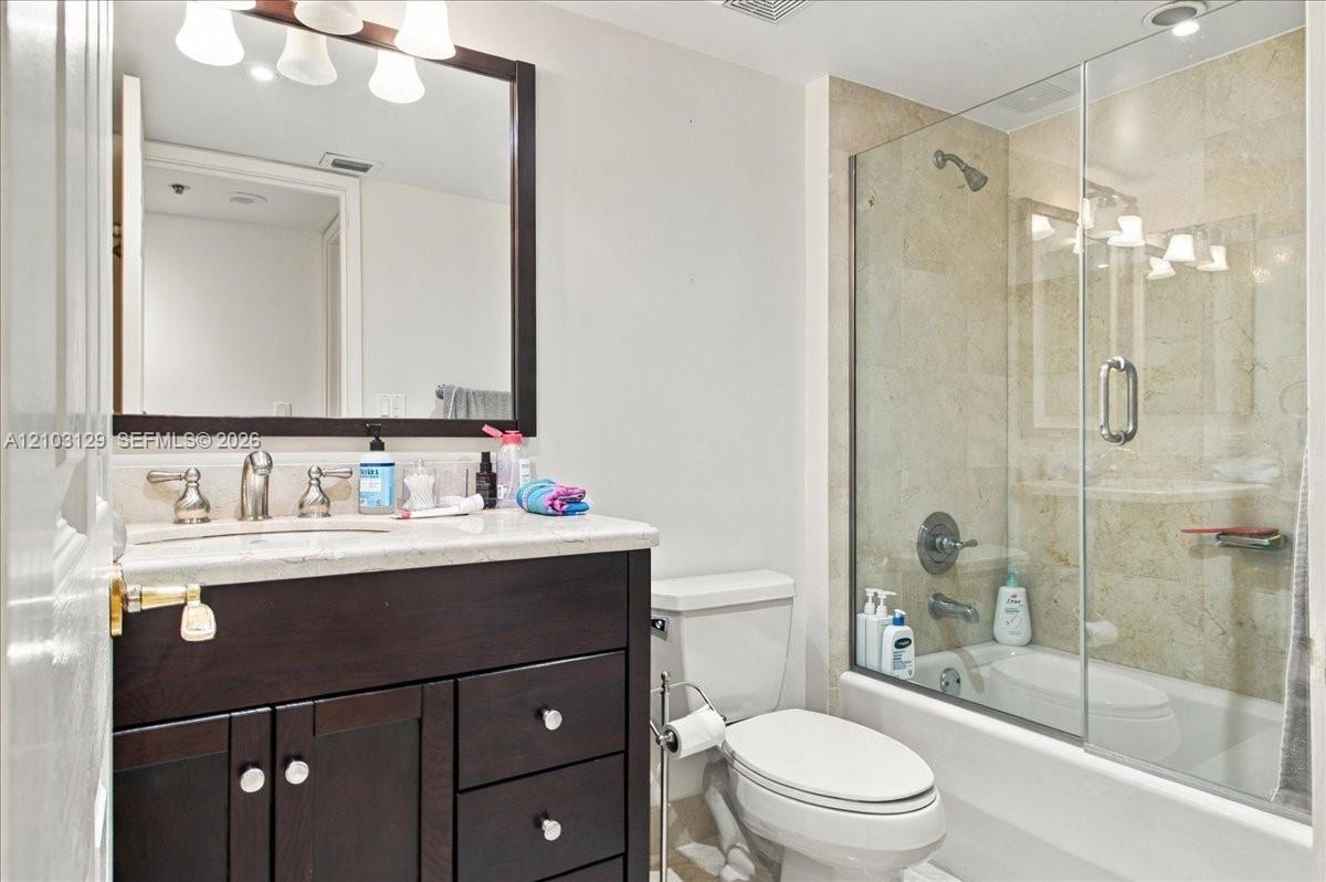Dark-finish wood cabinetry, stone-surface countertop, rectangular undermount sink, framed wall mirror, and tub-shower combination featuring a glass enclosure and stone-tile surround
