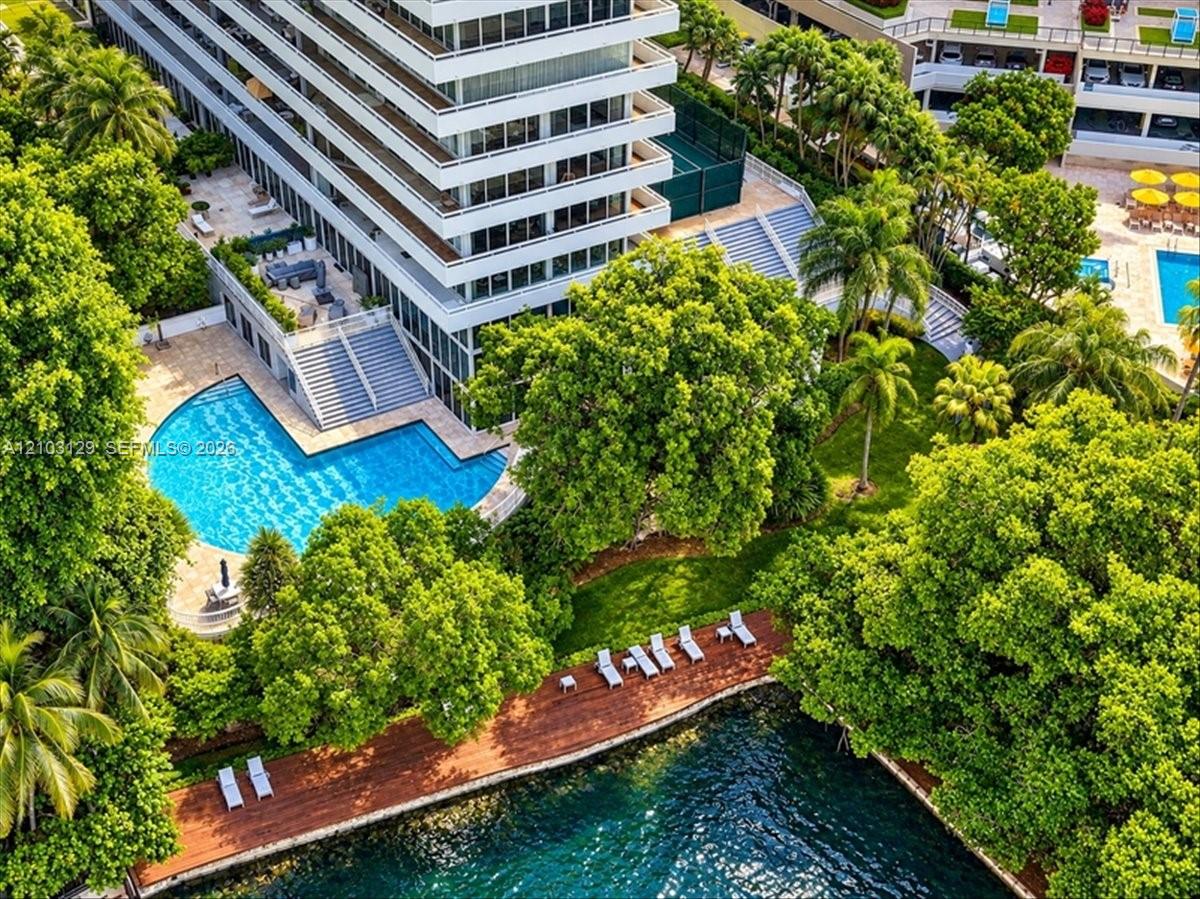 High-rise building exterior featuring tiered balconies, a sprawling swimming pool with surrounding terrace, a private tennis court, and a wood-decked shoreline