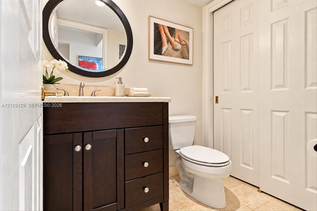 Dark wood vanity with a stone countertop, oval wall-mounted mirror, neutral stone flooring, and white two-panel closet doors