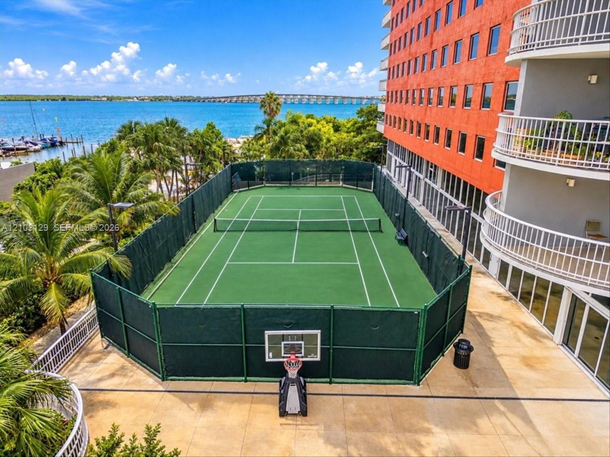 Tennis court enclosed by perimeter fencing, featuring a basketball hoop, set against a waterfront backdrop with established palm trees