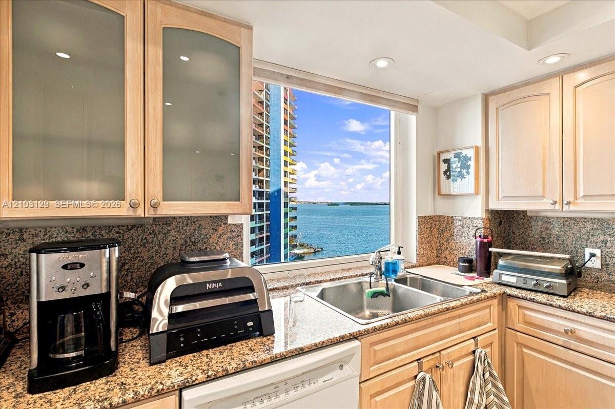 Kitchen featuring light wood cabinetry with frosted glass inserts, speckled stone countertops, dual-basin stainless steel sink, and recessed lighting