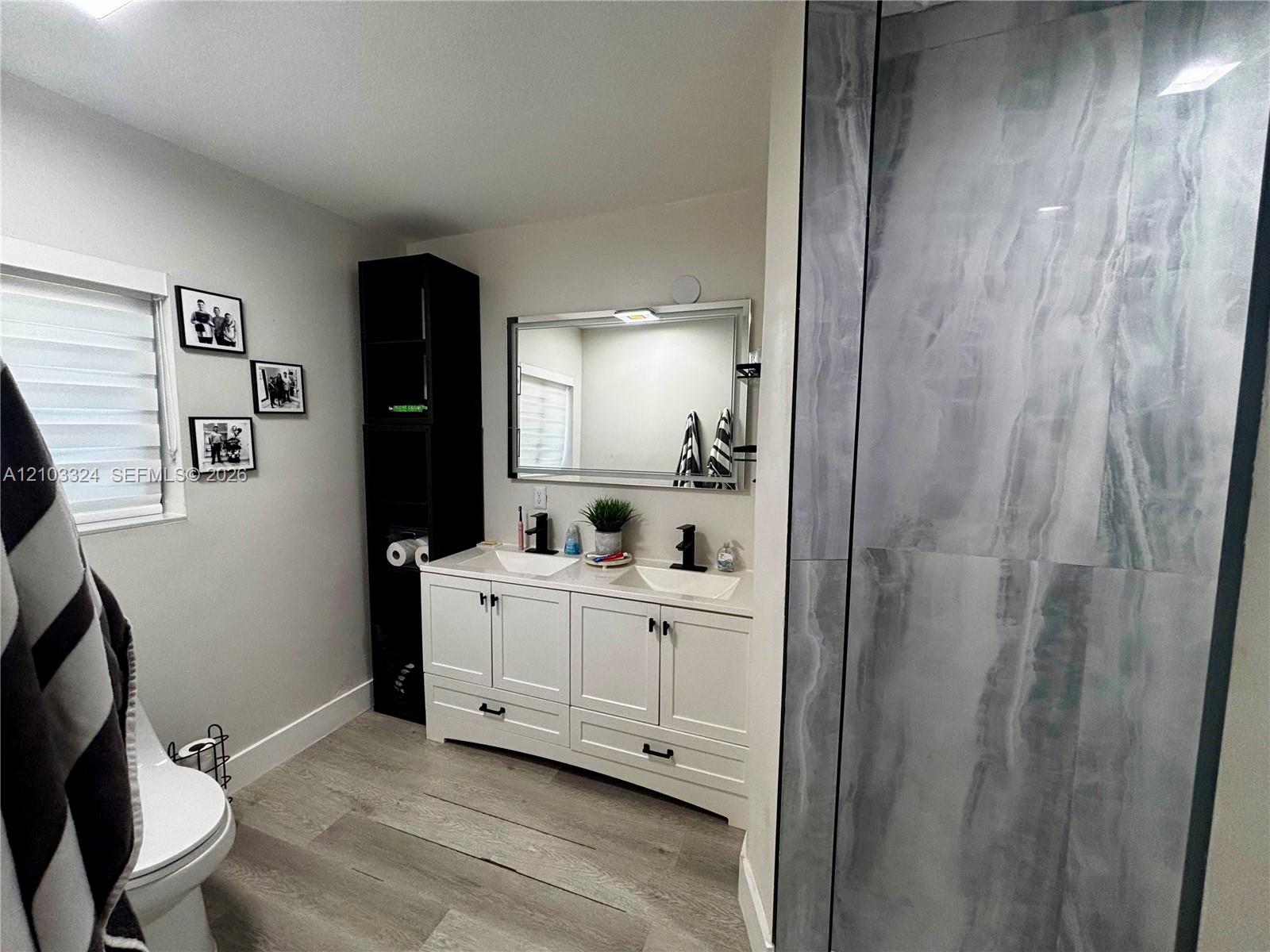 Dual-sink vanity with white cabinetry, wood-finish flooring, and walk-in shower featuring large-format tile surround