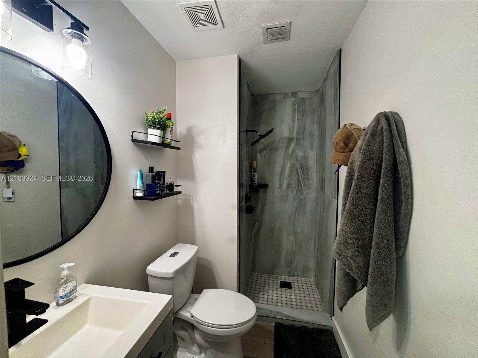 Bathroom featuring a vanity with a rectangular sink, matte black faucet, and circular mirror, complemented by a walk-in shower with floor-to-ceiling tile and built-in wall shelving
