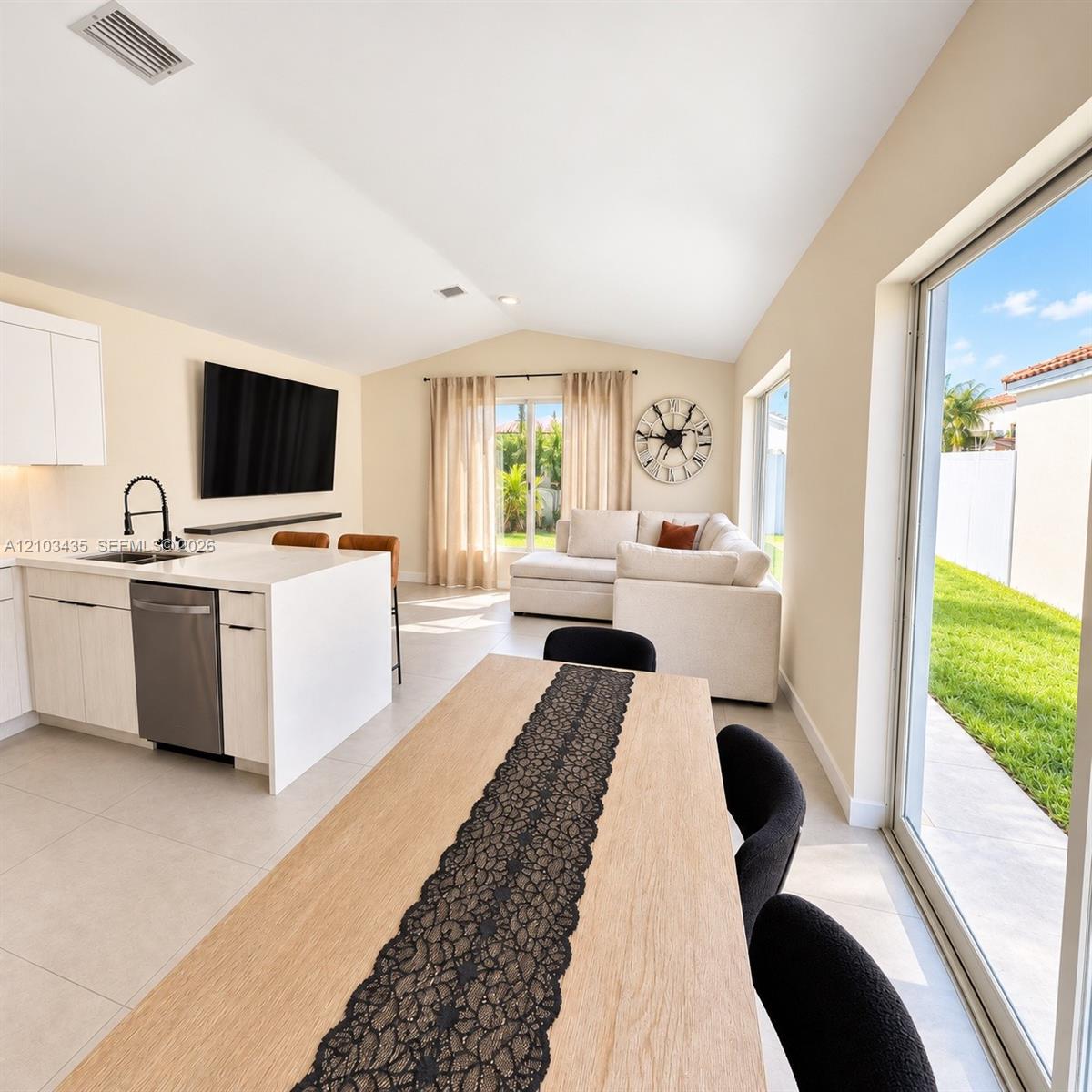 Open-concept interior featuring light-toned cabinetry, a stone-surface kitchen island with a matte black faucet, vaulted ceilings, and large sliding glass doors providing access to the exterior Open-concept interior featuring light-toned cabinetry, a stone-surface kitchen island with a matte black faucet, vaulted ceilings, and large sliding glass doors providing access to the exterior