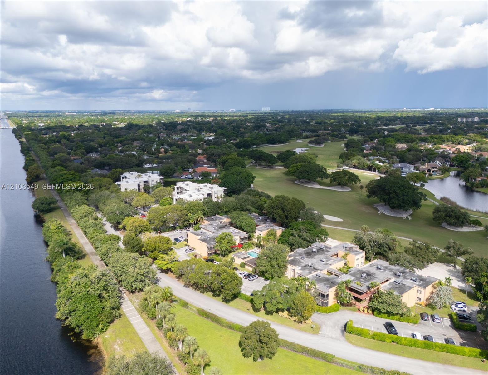 Aerial perspective of residential buildings situated along a waterway, featuring a community swimming pool, paved parking areas, and mature landscaping
