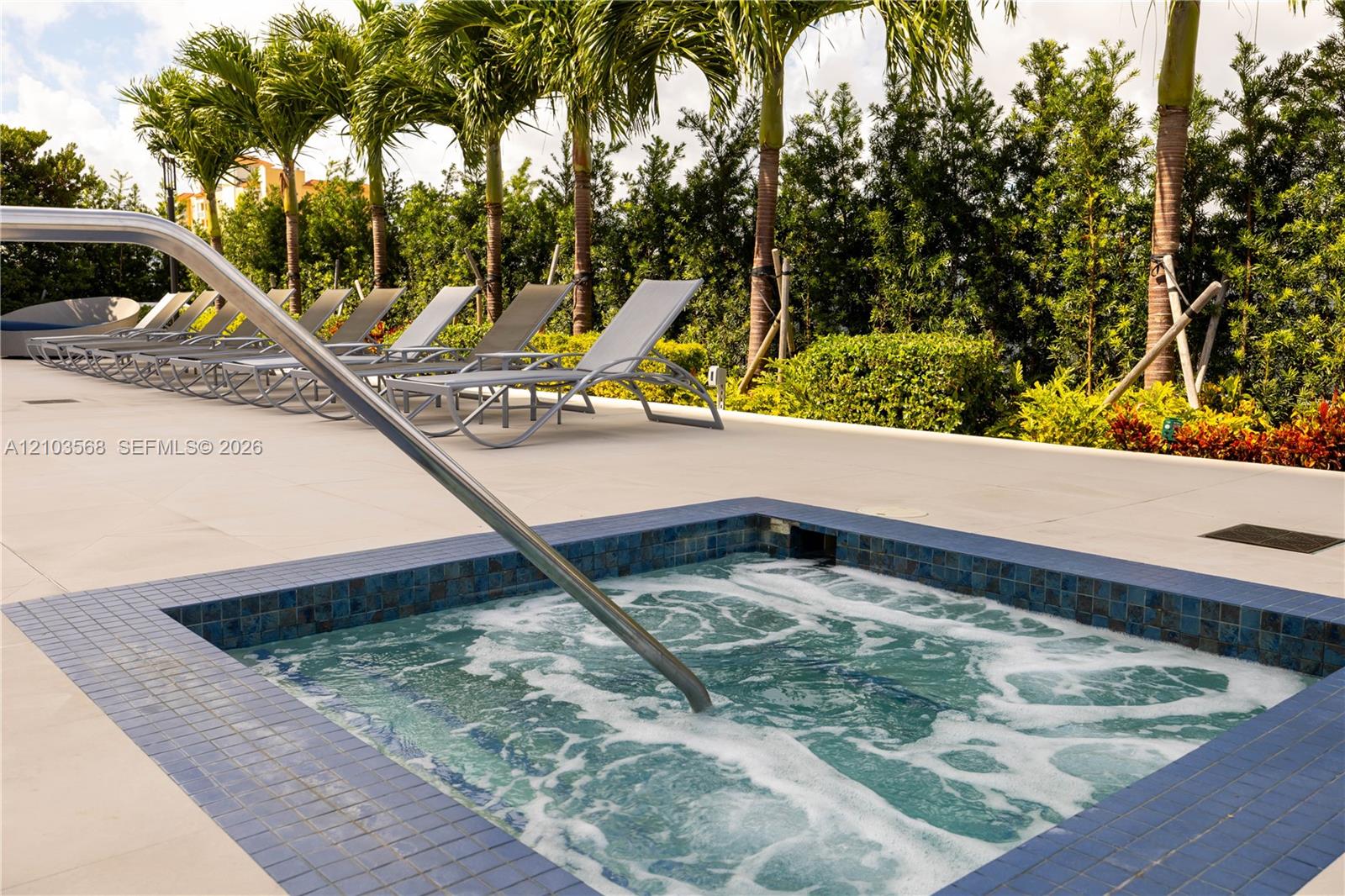 In-ground spa with blue tile surround, metal handrail, expansive concrete deck, and mature palm tree landscaping