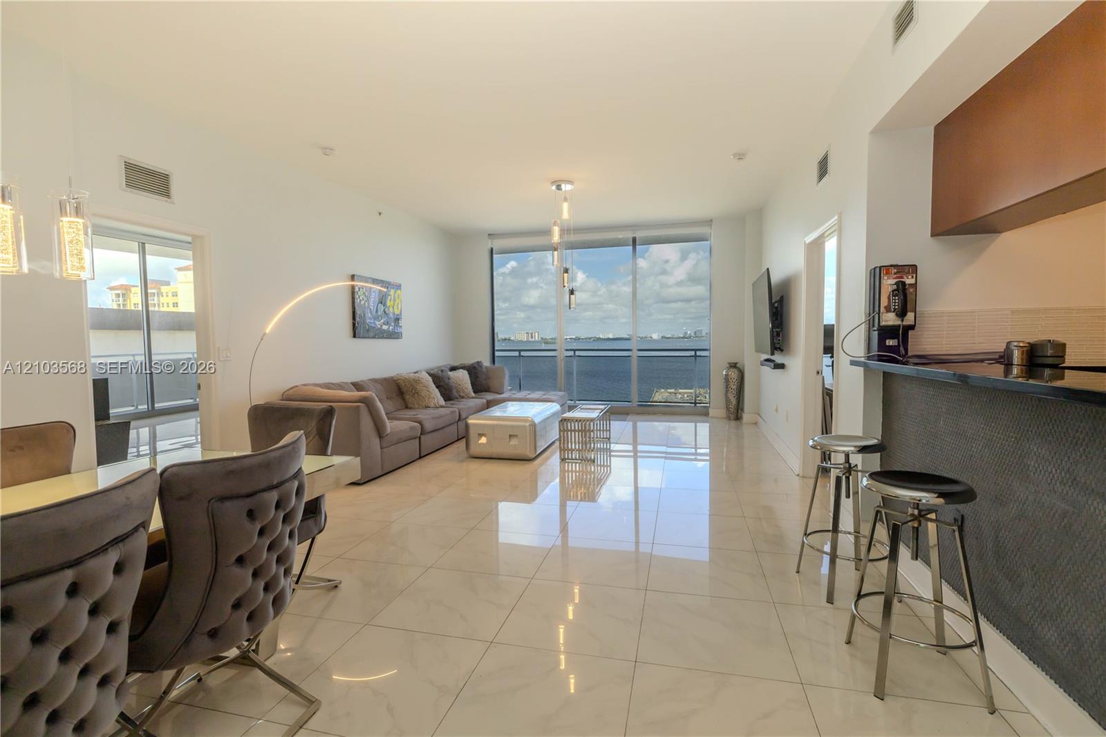 Open-concept living area featuring polished tile flooring, floor-to-ceiling windows, a stone-surfaced breakfast bar with dark finish cabinetry, and an attached balcony