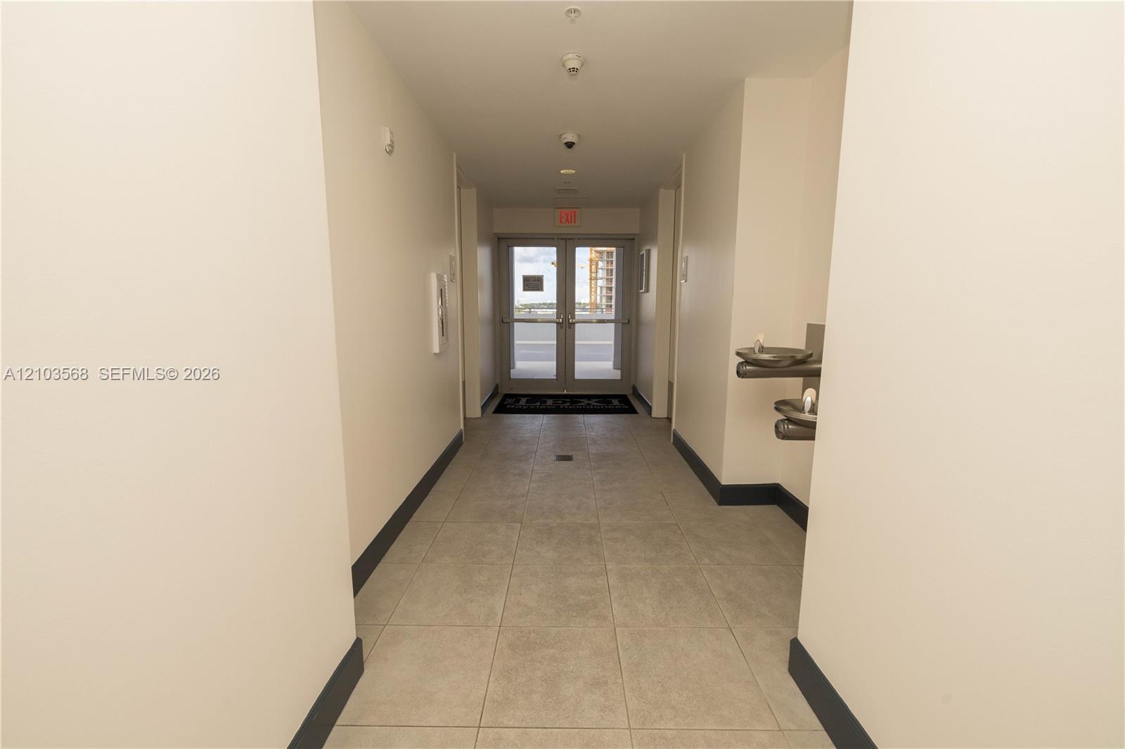 Interior hallway featuring neutral walls, large-format tile flooring, double glass-paneled doors, and wall-mounted metal water fountain station