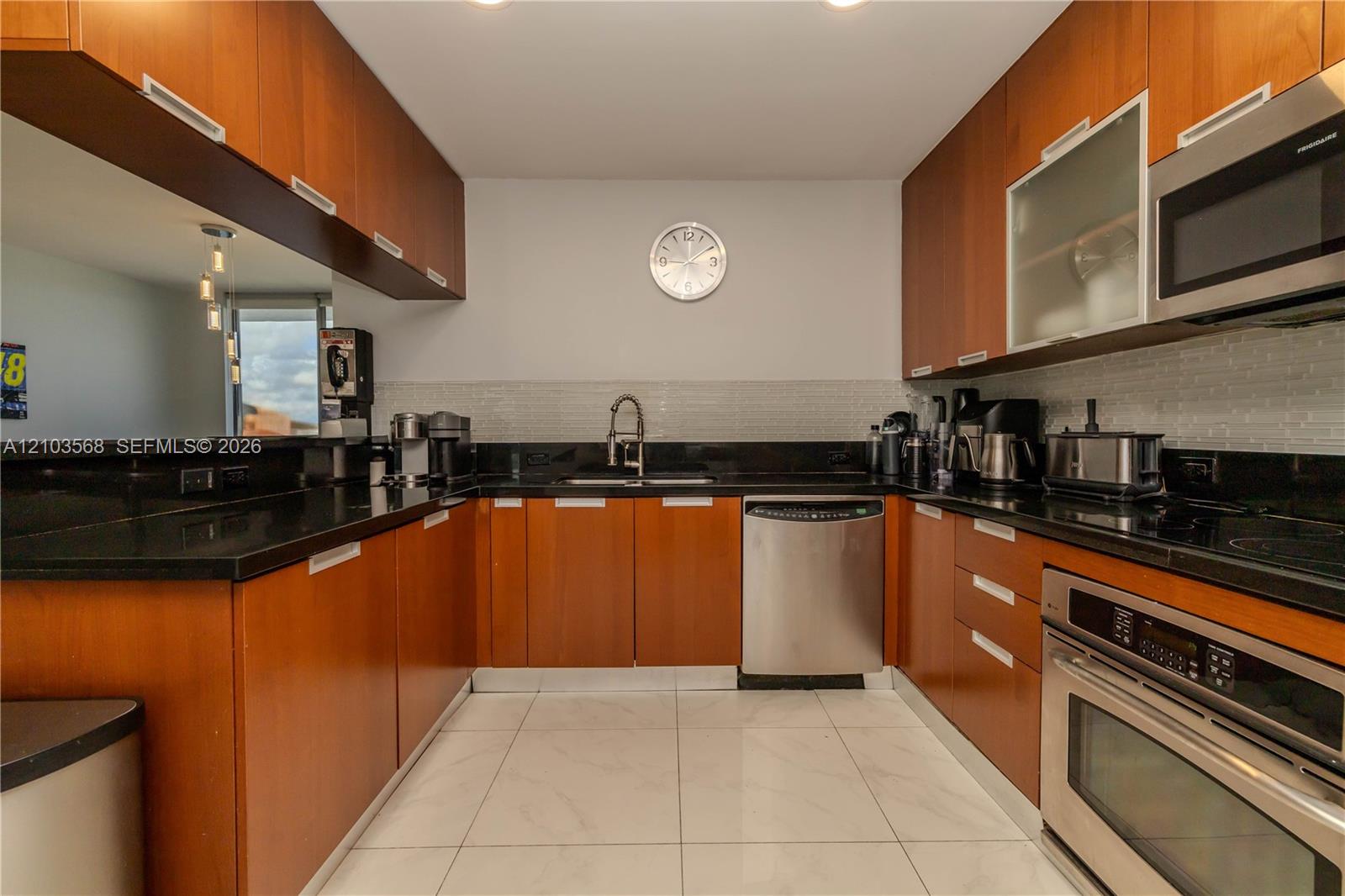U-shaped kitchen featuring wood-finish cabinetry, black stone countertops, stainless steel appliances, tiled backsplash, and light-toned tile flooring