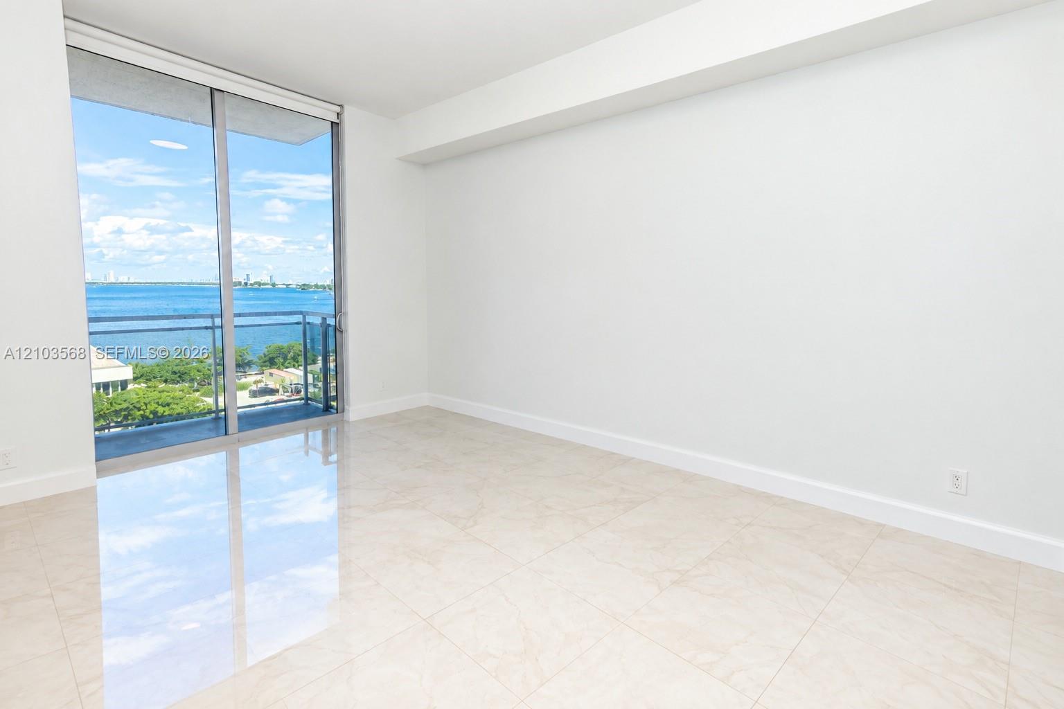 Floor-to-ceiling sliding glass doors, balcony access, neutral stone-finish tile flooring, and water-facing scenery