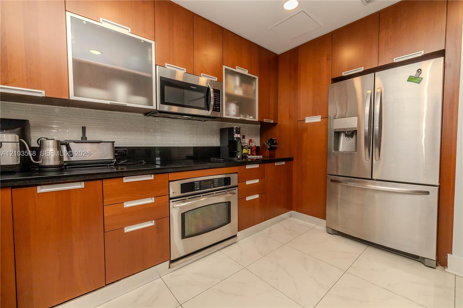 Kitchen featuring wood-finish cabinetry, stainless steel appliances, dark stone-style countertops, white subway tile backsplash, and large-format tile flooring