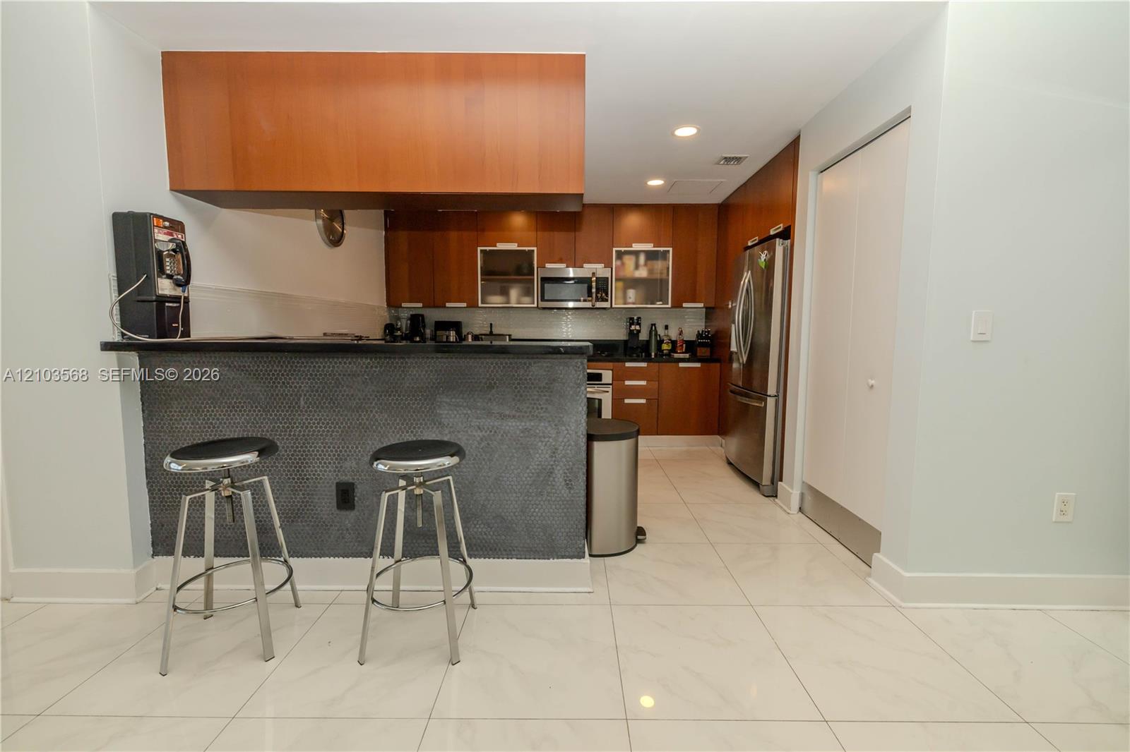 Kitchen featuring wood-finish cabinetry, a dark stone countertop, a textured breakfast bar, stainless steel appliances, and large-format tile flooring