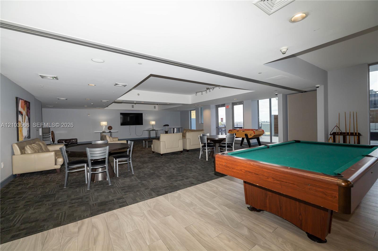 Expansive multi-use area featuring recessed ceiling details, wood-finish flooring, patterned carpeting, and floor-to-ceiling glass panel doors