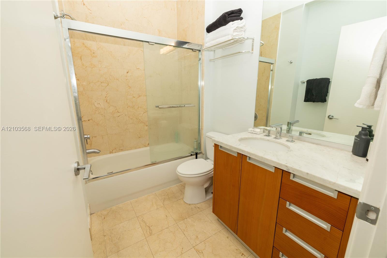 Full bathroom featuring stone-surround tub with glass enclosure, wood-finish vanity with stone countertop, undermount sink, and wall-to-wall mirror