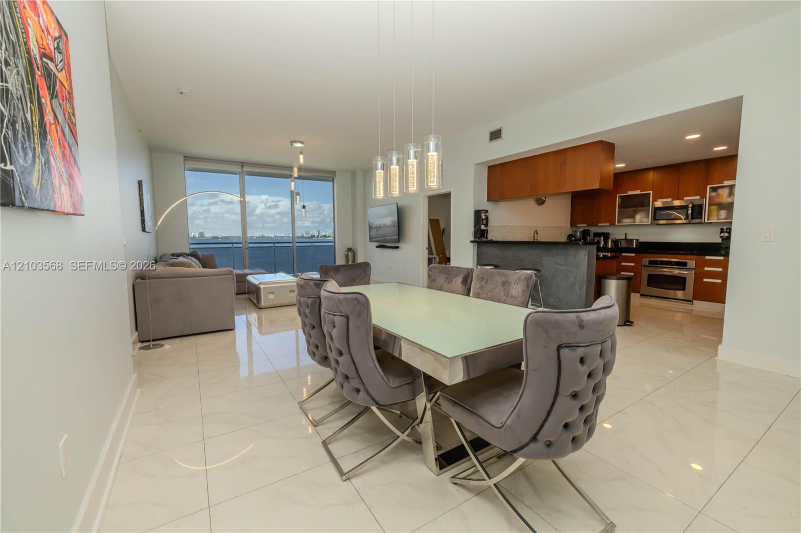 Open-concept floor plan featuring large-format polished tile flooring, floor-to-ceiling windows, built-in kitchen cabinetry with integrated stainless steel appliances, and a tiered granite-top breakfast bar