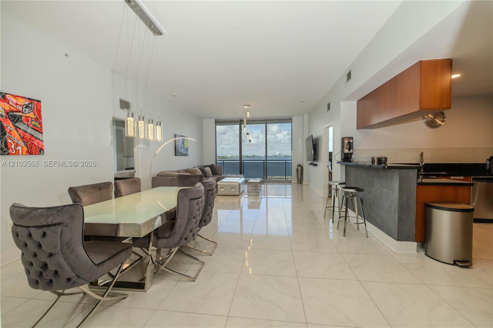 Open-concept interior featuring large-format tile flooring, a kitchen breakfast bar with stone-texture base, wood-finish upper cabinetry, a five-light linear pendant fixture, and floor-to-ceiling glass panel windows