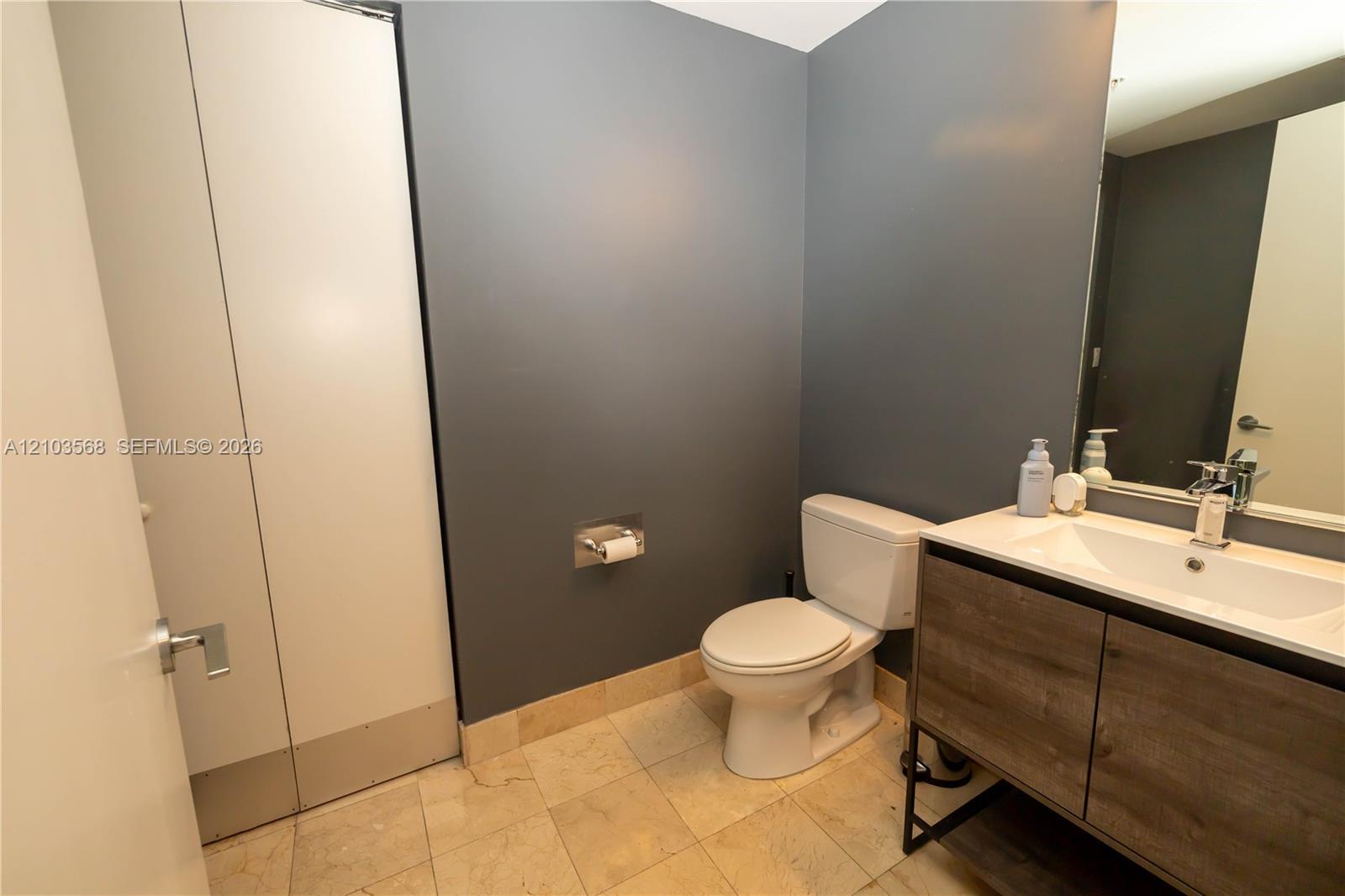 Modern vanity featuring a wood-finish cabinet with a white countertop and rectangular basin, paired with natural stone floor tiling and large-format wall mirror
