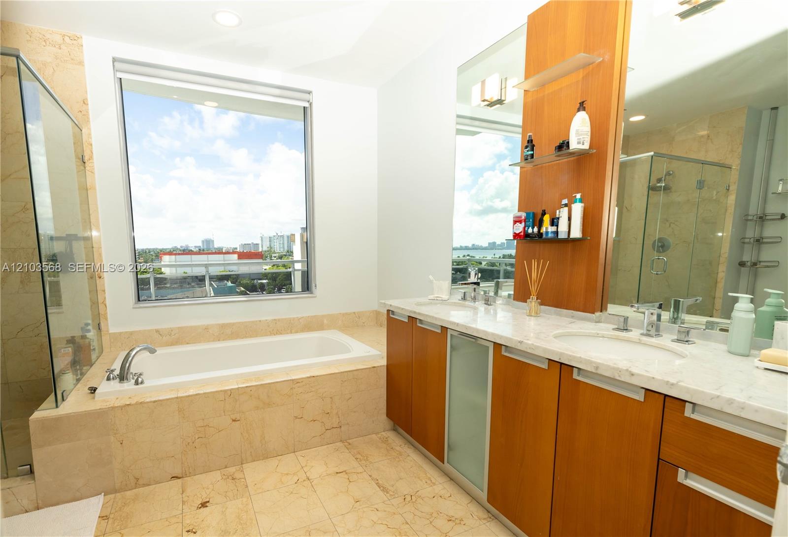 Bathroom featuring a deep soaking tub with a stone surround, dual-sink vanity with light-toned stone countertops, natural wood-finish cabinetry, a large frameless glass-enclosed shower, and expansive windows