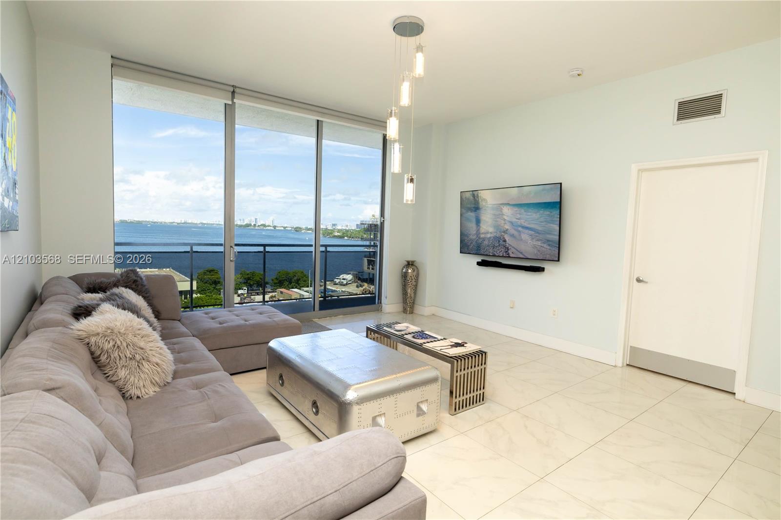Living space featuring floor-to-ceiling glass sliding doors, stone-finish tile flooring, a tiered multi-pendant light fixture, and a balcony with metal railings