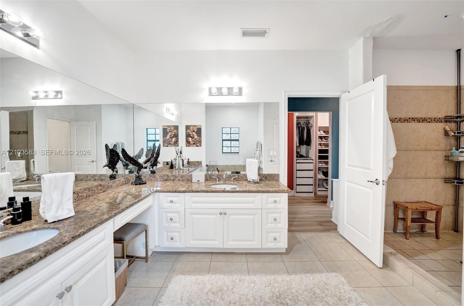 Dual vanity with white cabinetry, stone-finish countertops, expansive wall-to-wall mirror, and tiled shower enclosure