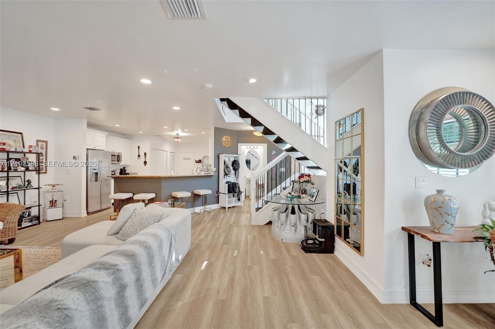 Open-concept interior featuring light-toned wood-finish flooring, recessed lighting, a kitchen breakfast bar with stone-finish countertops, and an architectural staircase with white railings and dark-toned treads