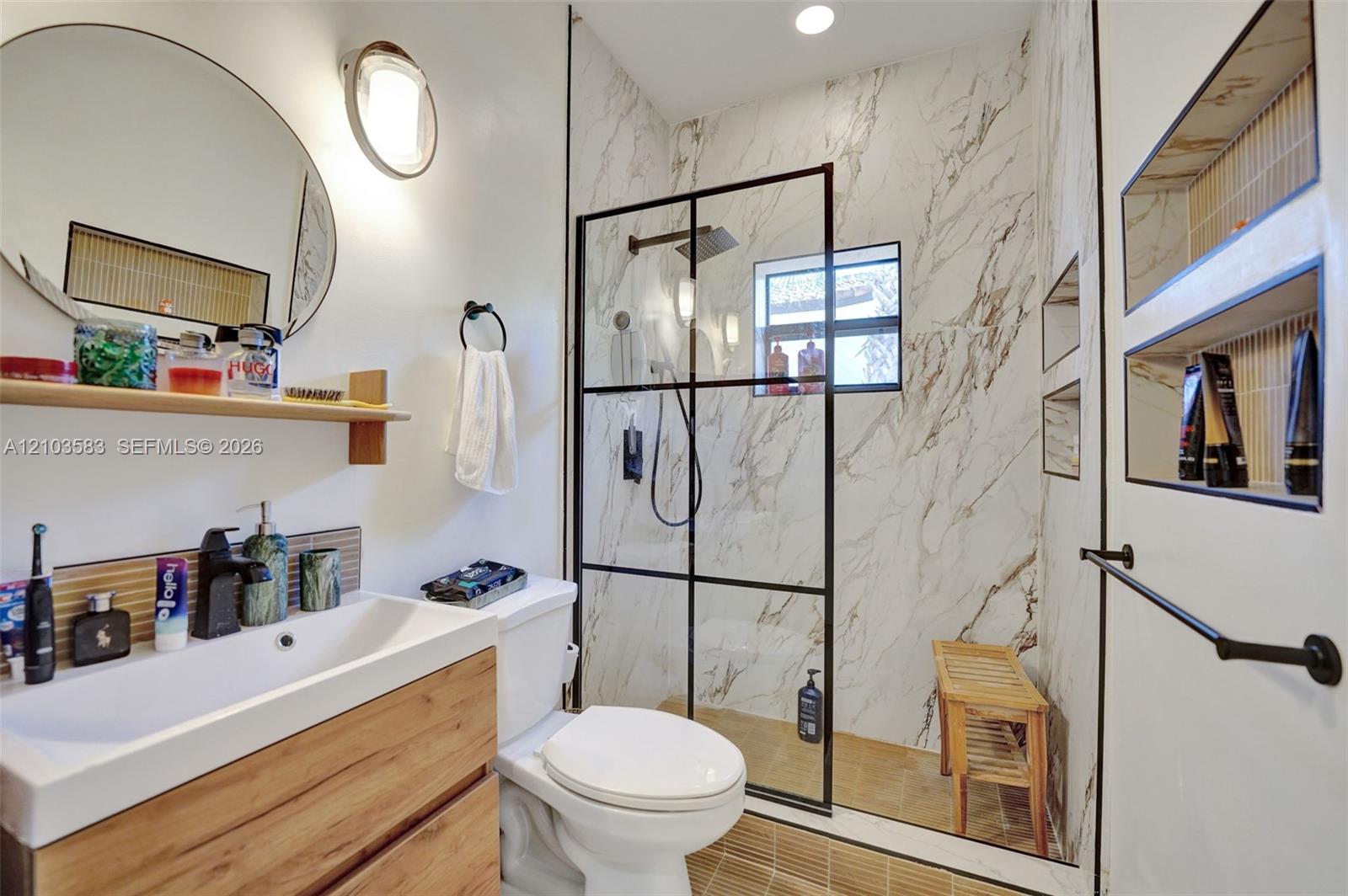 Wood-finish floating vanity with a white rectangular sink, walk-in shower with black-framed glass enclosure, stone-tiled shower walls, matte black fixtures, and recessed wall shelving