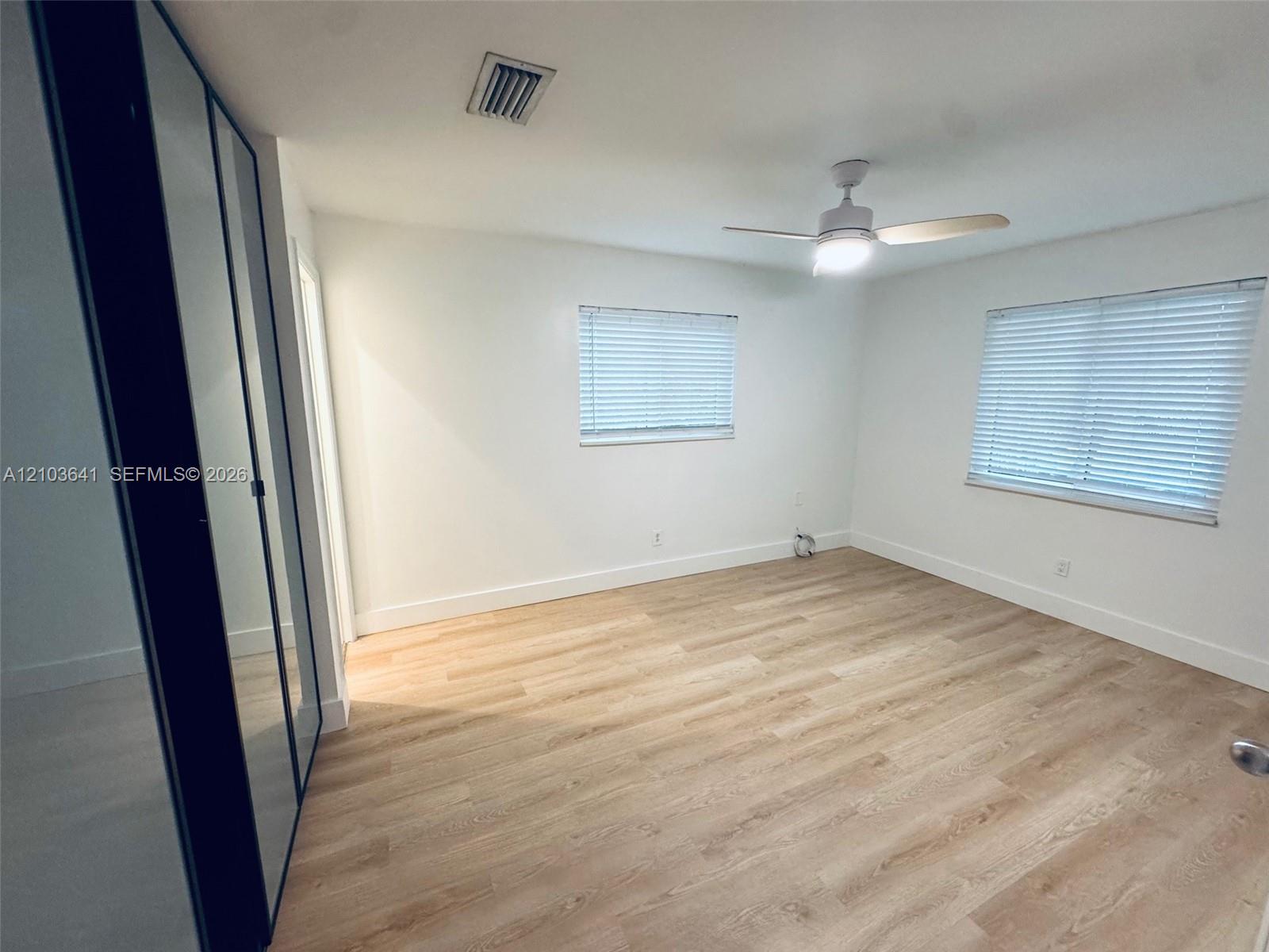 Light-filled room featuring wood-finish flooring, dual windows with blinds, a ceiling fan with integrated lighting, and full-length sliding mirror-paneled closet doors