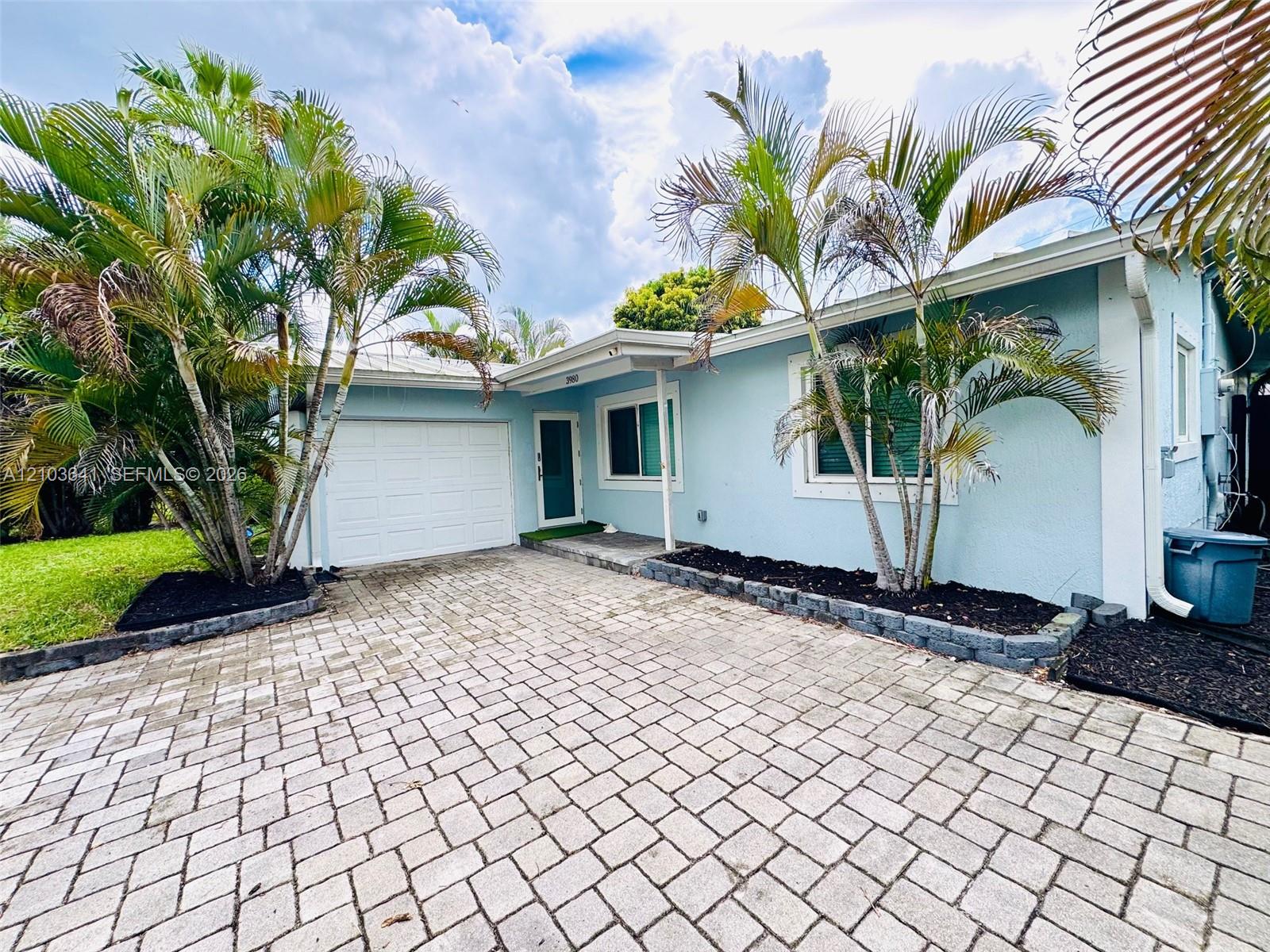 Single-story exterior with light blue stucco siding, paver driveway, single-car garage, and integrated landscaping featuring mature palms and stone-bordered garden beds