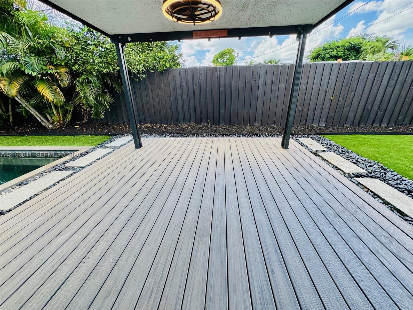 Covered outdoor deck featuring composite decking, a built-in overhead light fixture, stone-lined paver borders, and artificial turf surroundings