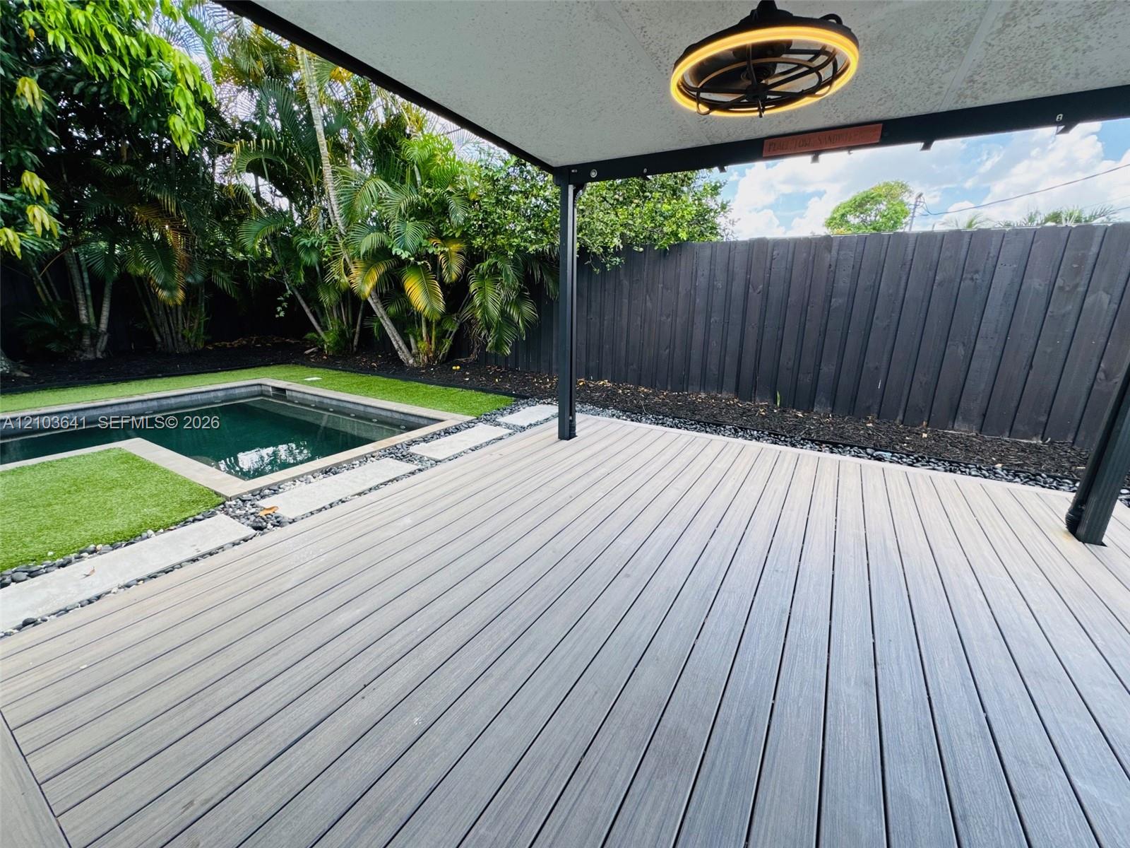 Covered deck with composite wood-finish planking, recessed ceiling lighting, fenced backyard, and inground pool with synthetic turf surround
