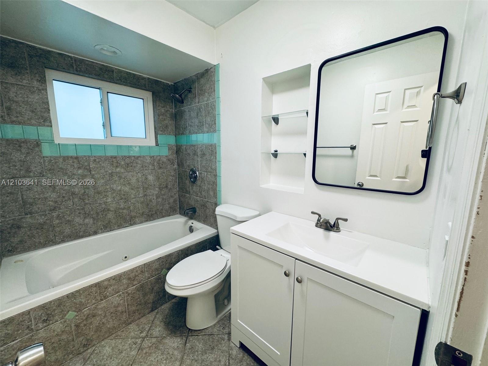 Bathroom featuring a white vanity cabinet with an integrated sink, rectangular mirror with a black frame, recessed wall shelving, and a combination tub and shower with gray tile surround and turquoise accent tiling