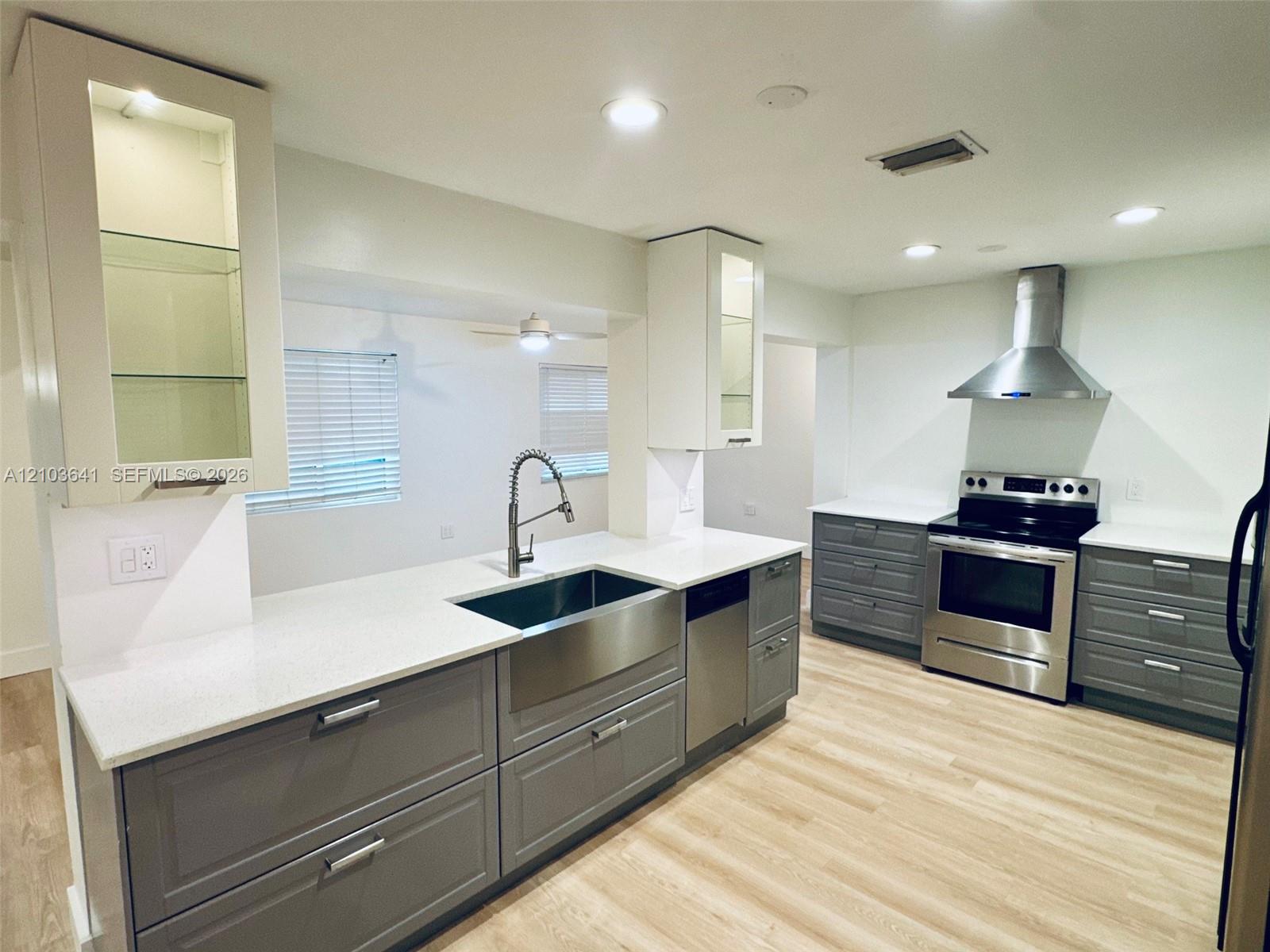 Kitchen featuring white quartz countertops, grey shaker-style cabinetry, stainless steel farmhouse-style sink, stainless steel range hood, and wood-finish flooring