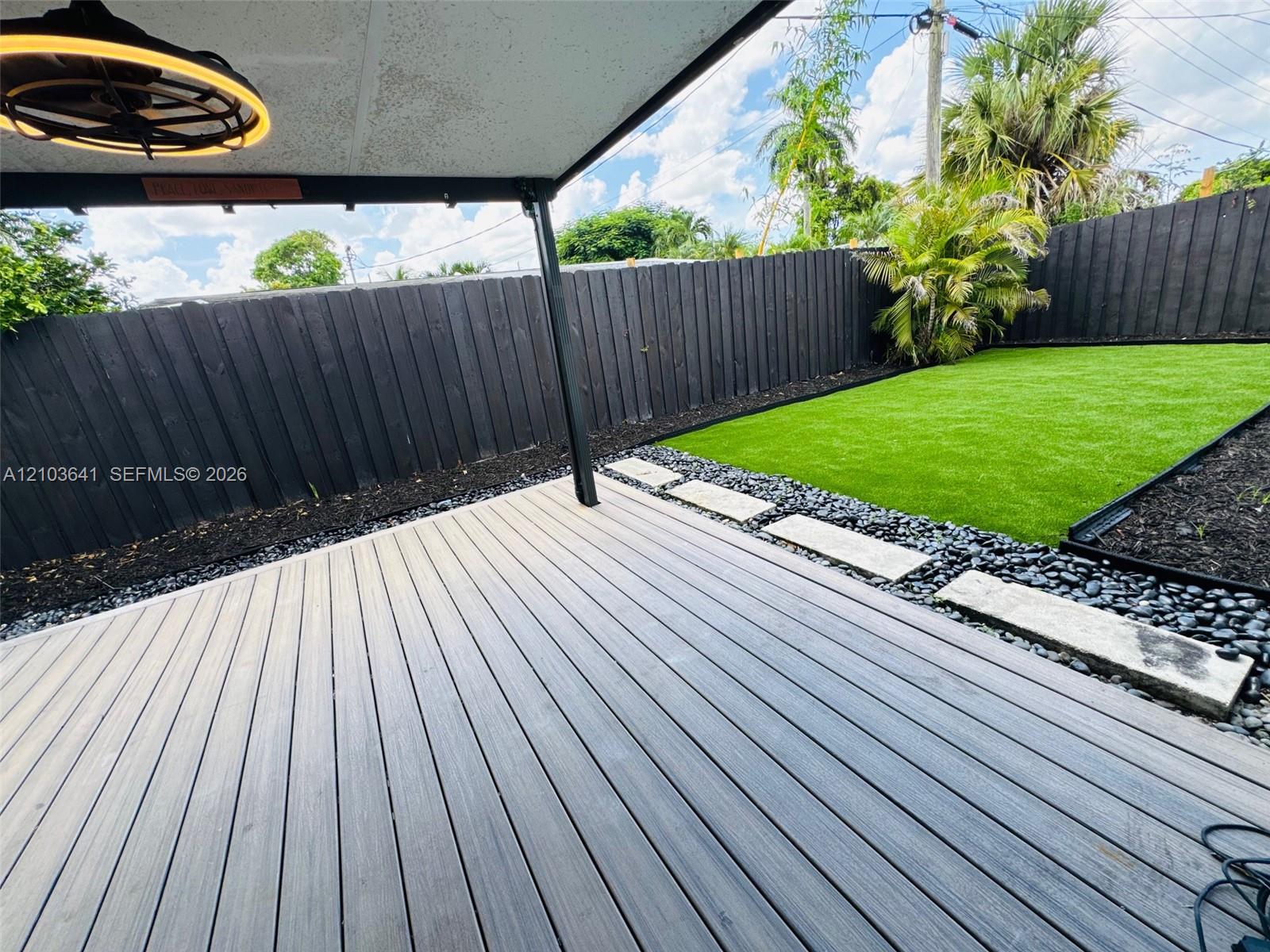 Covered patio featuring composite decking, synthetic turf lawn, stone-accented border, and a black privacy fence